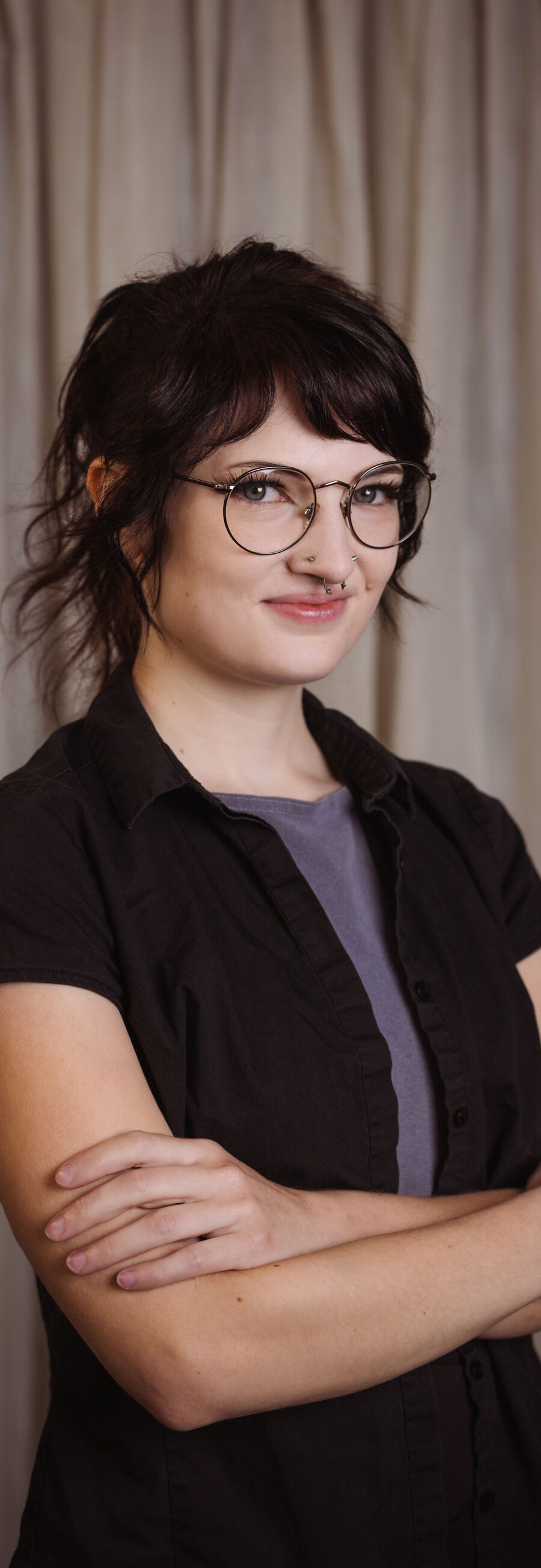 Grace, smiling with arms crossed, wearing glasses and a black shirt in front of a studio curtain backdrop