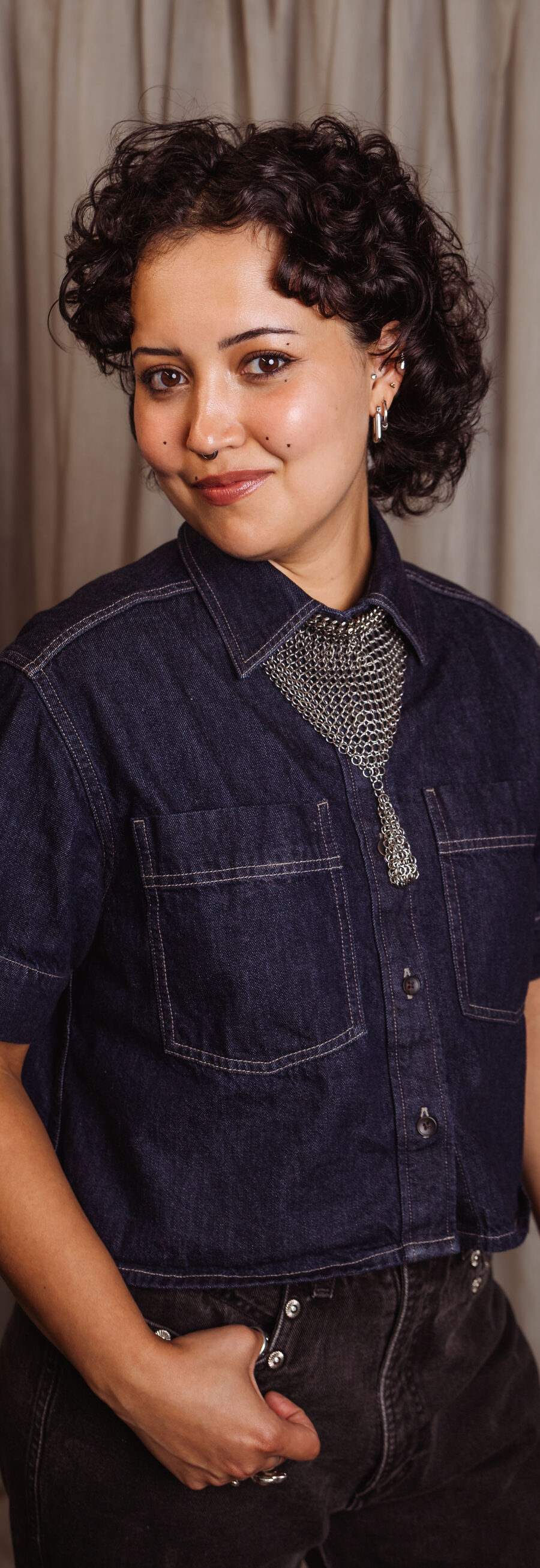 Morgan, smiling, with short dark curls, in a dark denim shirt in front of a studio curtain backdrop