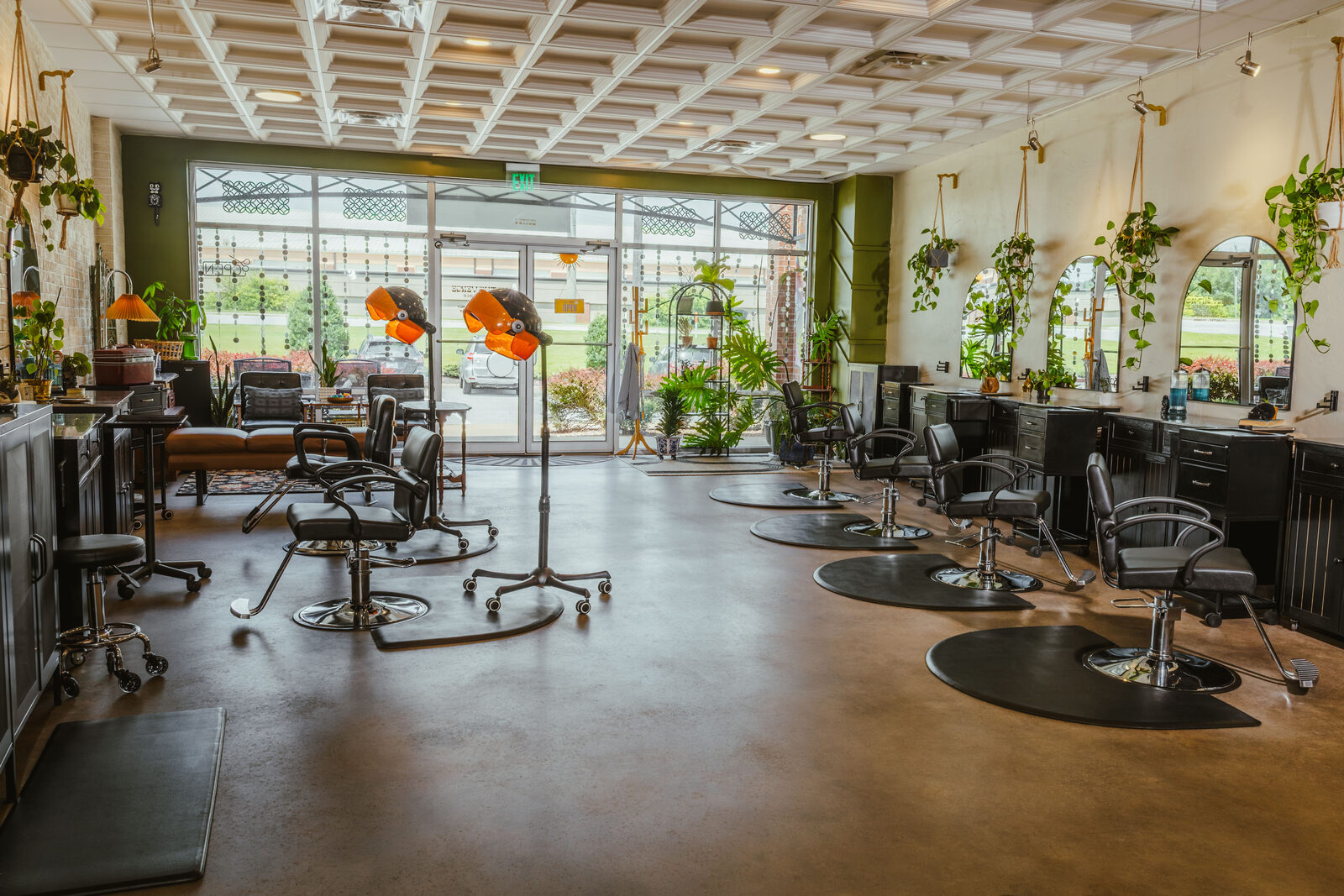 Inside On The Verge salon: black styling chairs and vintage orange hooded dryers on a polished concrete floor, with hanging plants, arched mirrors and a sunlit glass storefront
