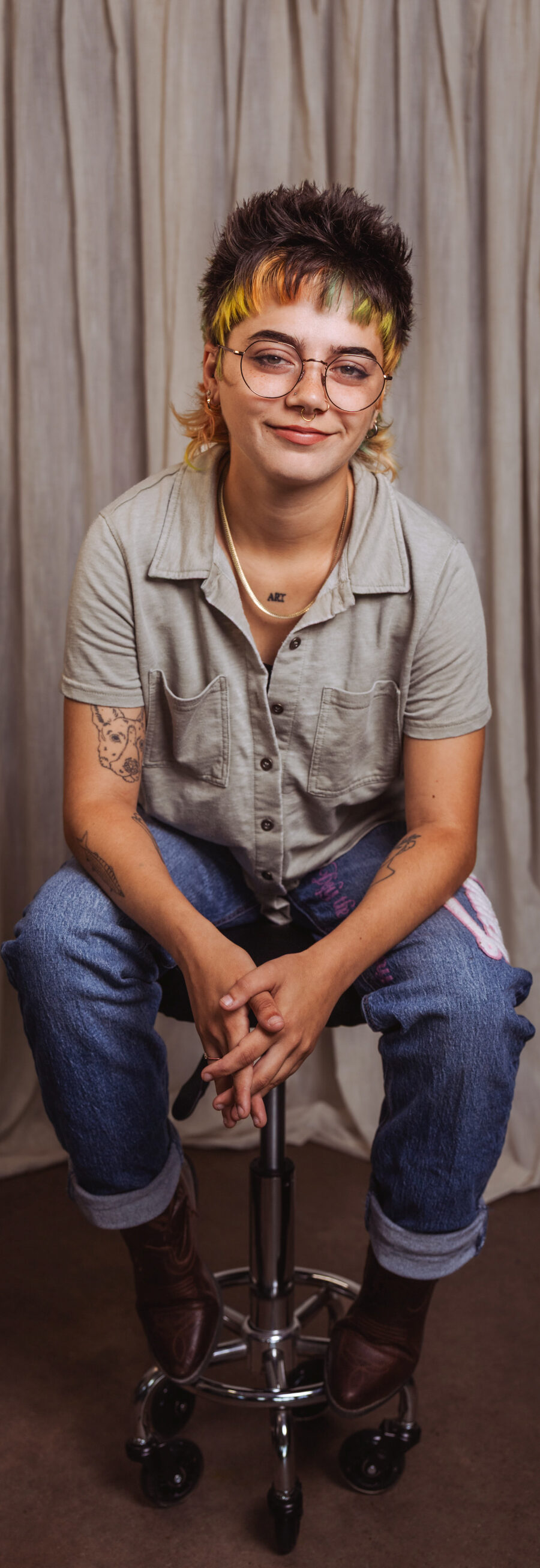 Taylor, smiling, seated on a stool in rolled-up jeans, with glasses and a yellow-fringed mullet, in front of a studio curtain backdrop