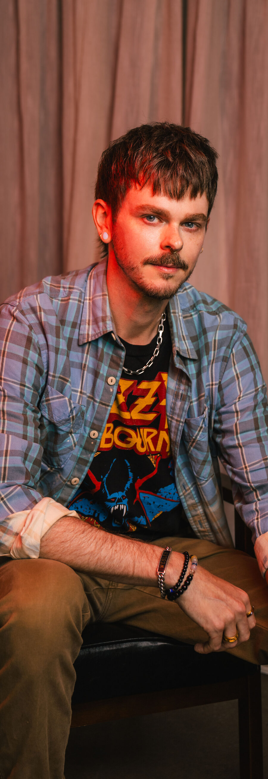 Wes, seated in a leather chair, wearing a plaid shirt over a band tee, in front of a studio curtain backdrop